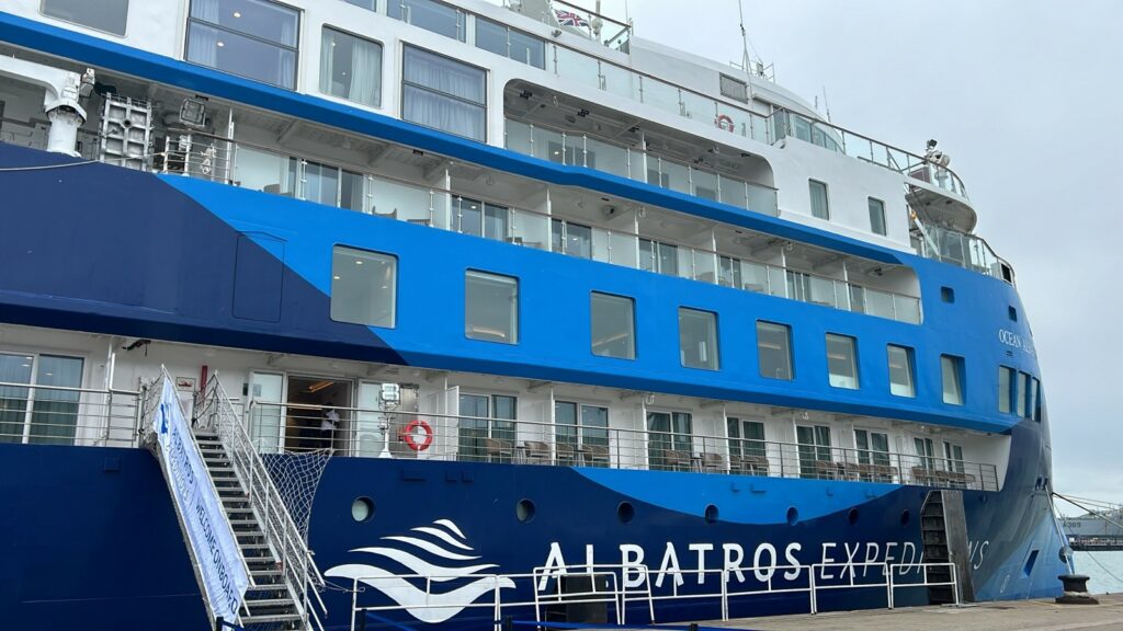 Außenansicht des Kreuzfahrtschiffes "Ocean Albatros". Man erkennt einige Fenster der Kabinen an Bord, mit Balkonen auf denen jeweils Tische und Stühle stehen. Das Schiff hat eine Lackierung in verschiedenen Blautönen. 