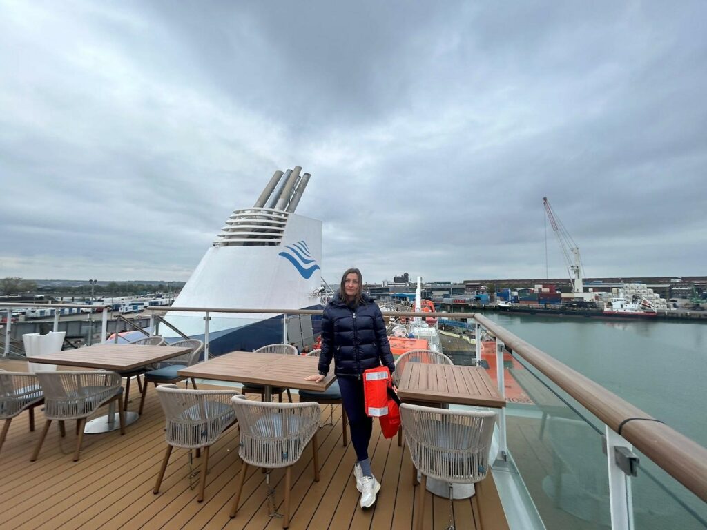 Frau steht an Bord eines Schiffes. Im Hintergrund ist der Hafen zu erkennen, sowie die Trichter des Schiffes. Die Frau steht zwischen Stühlen und Tischen an Deck. Der Himmel ist grau und bewölt. 