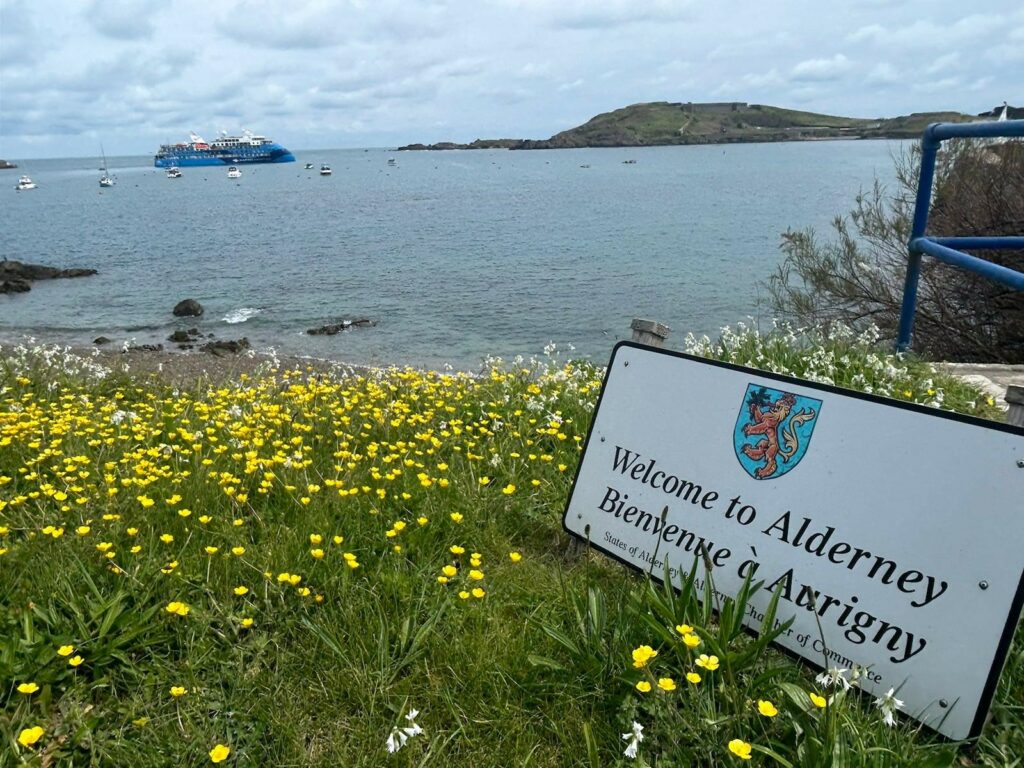 Im Vordergrund erkennen wir eine kleine Wiese mit gelben Blumen. An der rechten Bildseite sehen wir ein weißes Schild mit Wappen der Insel Alderney, sowie schwarzem Schriftzug. Der Schriftzug lautet "Welcome to Alterney" zu Deutsch "Willkommen in Alderney". 
Im Hintergrund sehen wir erneut im Wasser das Kreuzfahrtschiff Ocean Albatros, kleinere Boote sowie eine Landzunge die ins Wasser ragt. 